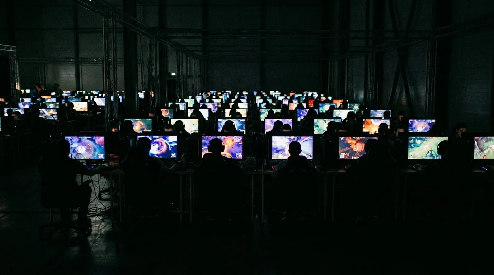 A near-black gaming club room where the only light falls on the monitors and keyboards of the stations